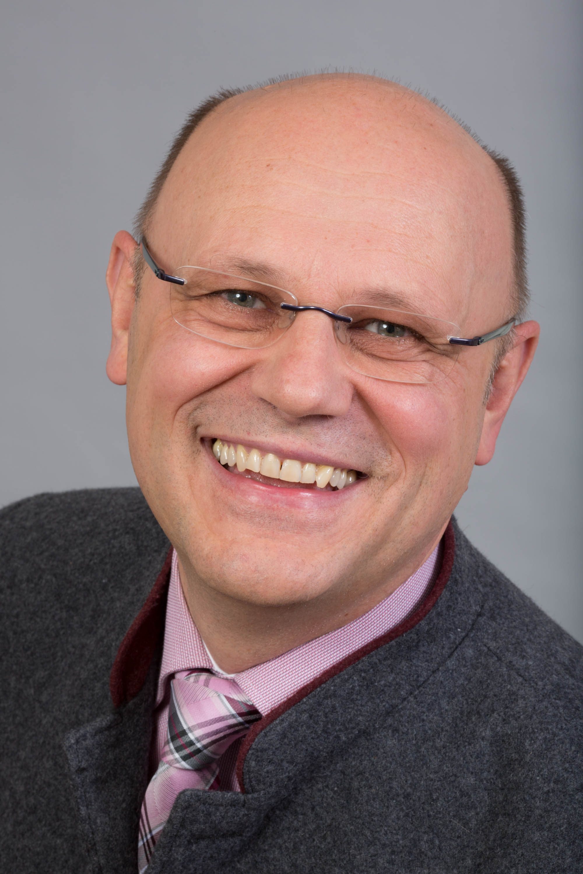Ein Mann mittleren Alters mit Brille, Glatze und einem breiten Lächeln trägt einen grauen Blazer und ein rosa kariertes Hemd mit gestreifter Krawatte und präsentiert sich als selbstbewusster Organisationsleiter vor einem schlichten grauen Hintergrund.