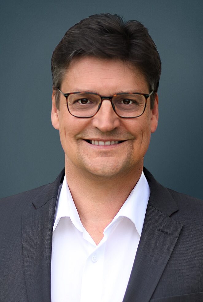 Ein Mann mit kurzen braunen Haaren und Brille lächelt in die Kamera. Er trägt eine dunkle Anzugsjacke über einem weißen Hemd mit Kragen und hat ein professionelles Erscheinungsbild, das seine Rolle in der Organisation widerspiegelt, und steht vor einem schlichten dunklen Hintergrund.
