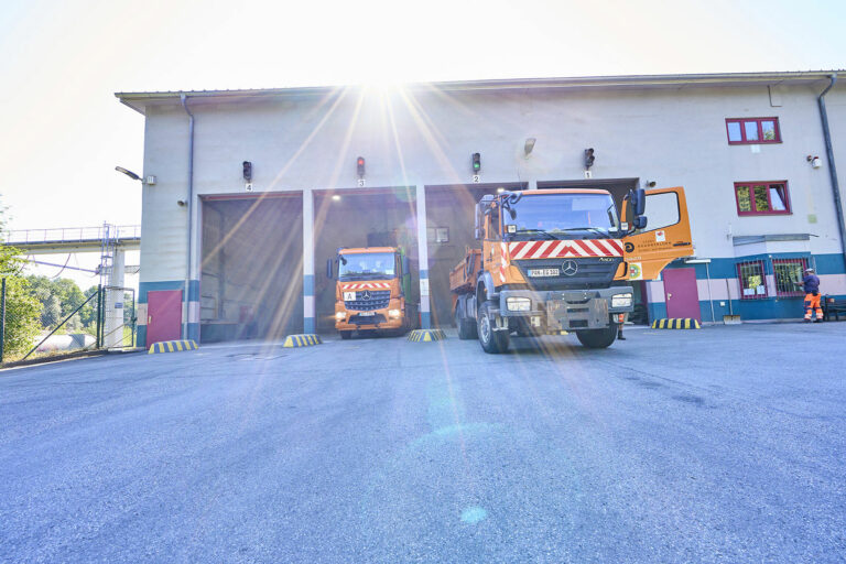 Zwei orangefarbene, gestreifte Nutzfahrzeuge fahren bei hellem Sonnenlicht aus einem Gebäude heraus. Die Garagentore sind geöffnet, und ein Arbeiter in orangefarbener Uniform steht rechts neben dem Gebäude.