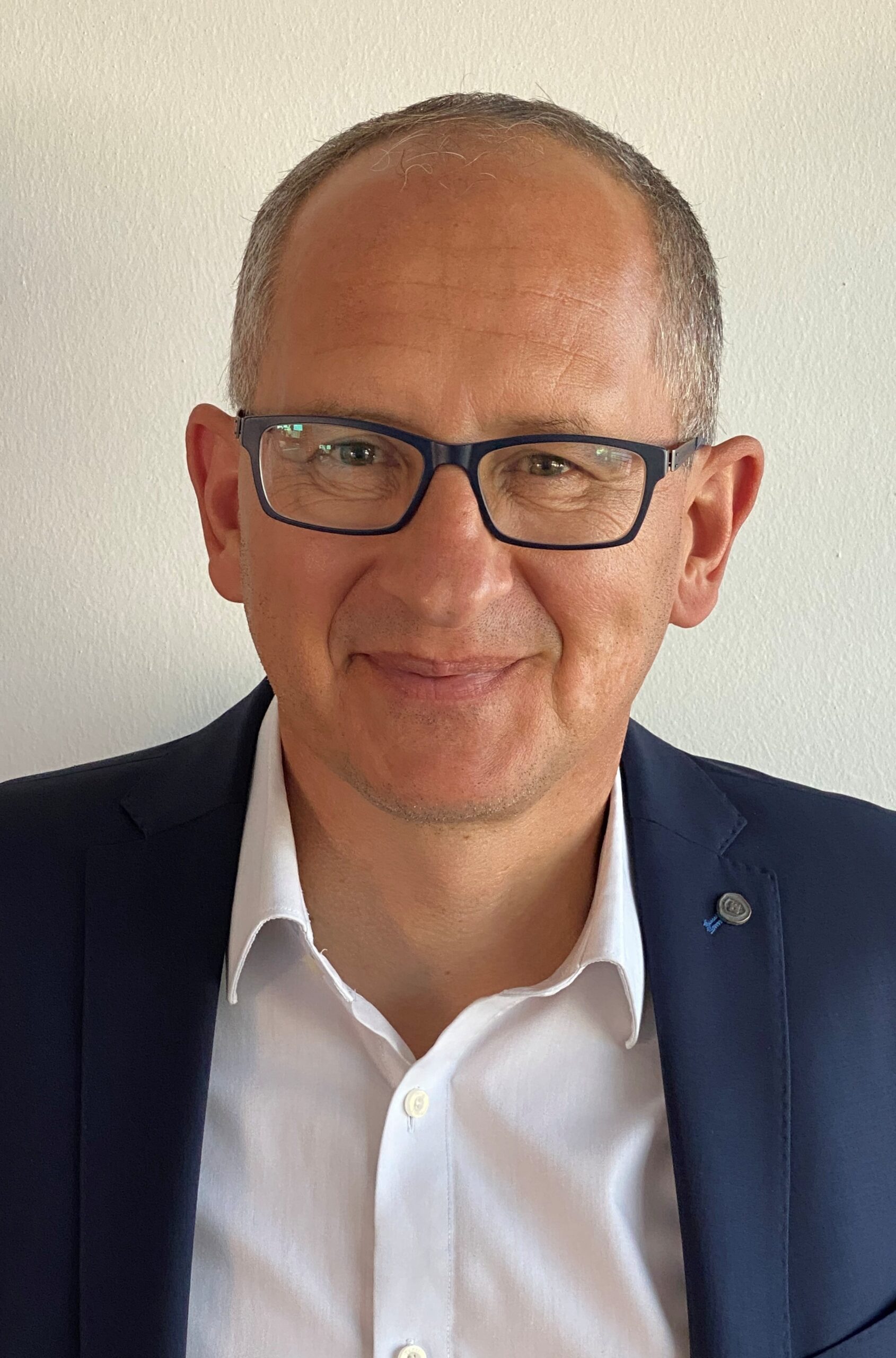 Ein Mann mittleren Alters mit kurzen Haaren und Brille, der einen dunklen Blazer und ein weißes Hemd trägt, lächelt in die Kamera vor einem schlichten hellen Hintergrund.