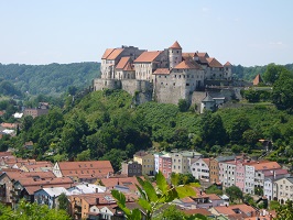 Eine große mittelalterliche Burg mit roten Dächern thront auf einem grünen Hügel im Verbandsgebiet und überblickt eine Stadt mit bunten Häusern und Bäumen unter einem klaren blauen Himmel.
