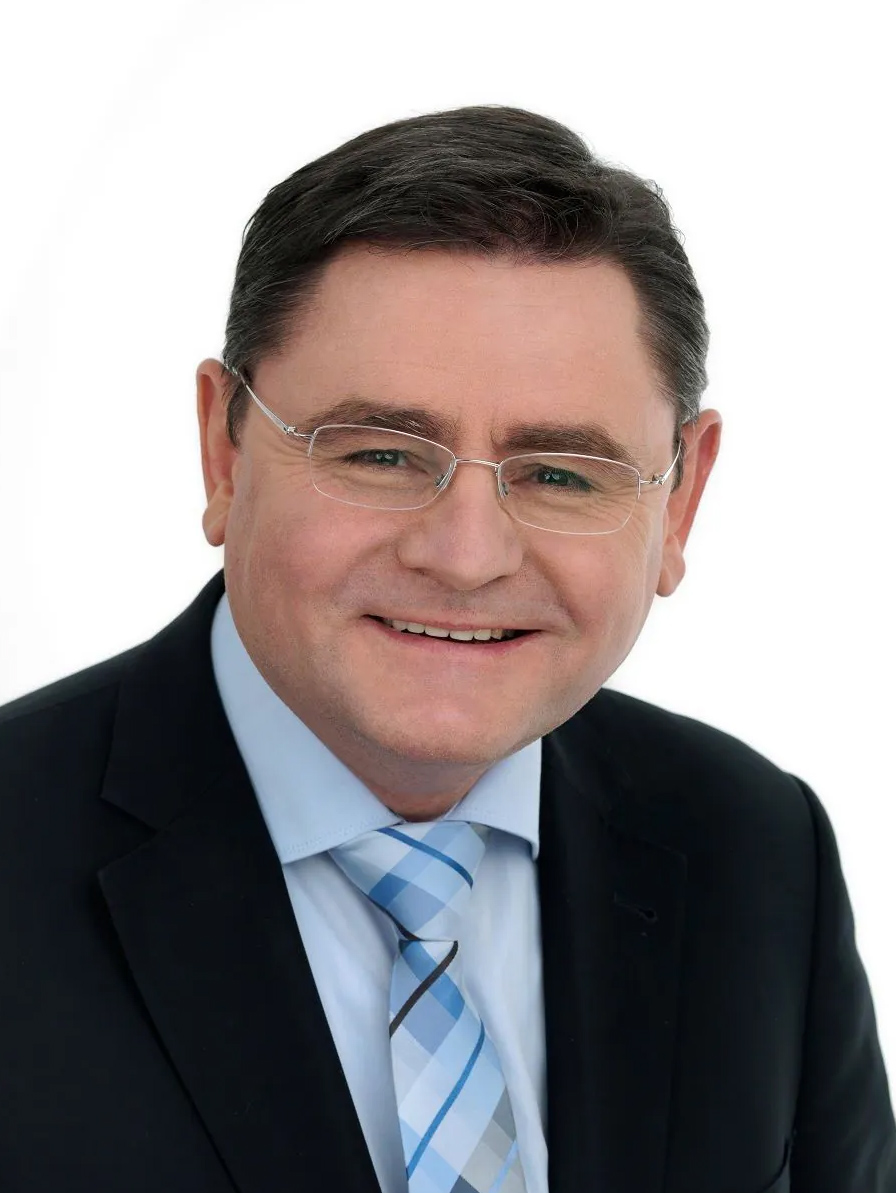 Ein Mann mittleren Alters mit kurzen dunklen Haaren, der eine Brille, einen dunklen Anzug, ein hellblaues Hemd und eine blau gemusterte Krawatte trägt, lächelt vor einem schlichten weißen Hintergrund in die Kamera und repräsentiert die Organisation ZAS.