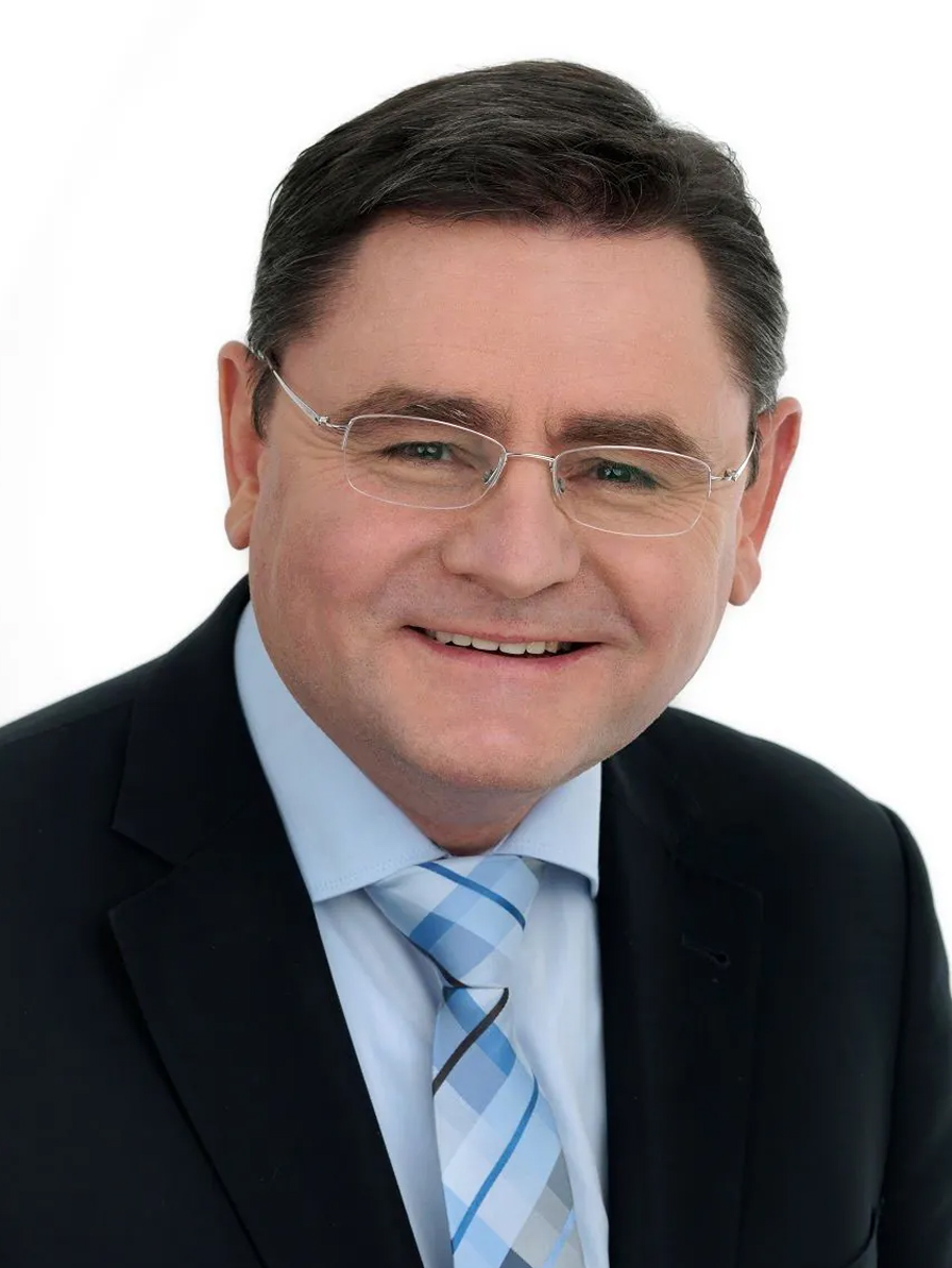 Ein Mann in Anzug und Krawatte lächelt in die Kamera. Er vertritt die ZAS-Organisation, hat kurze dunkle Haare, trägt eine Brille und ist mit einem hellblauen Hemd und einer gemusterten blau-grauen Krawatte bekleidet. Der Hintergrund ist einfarbig weiß.