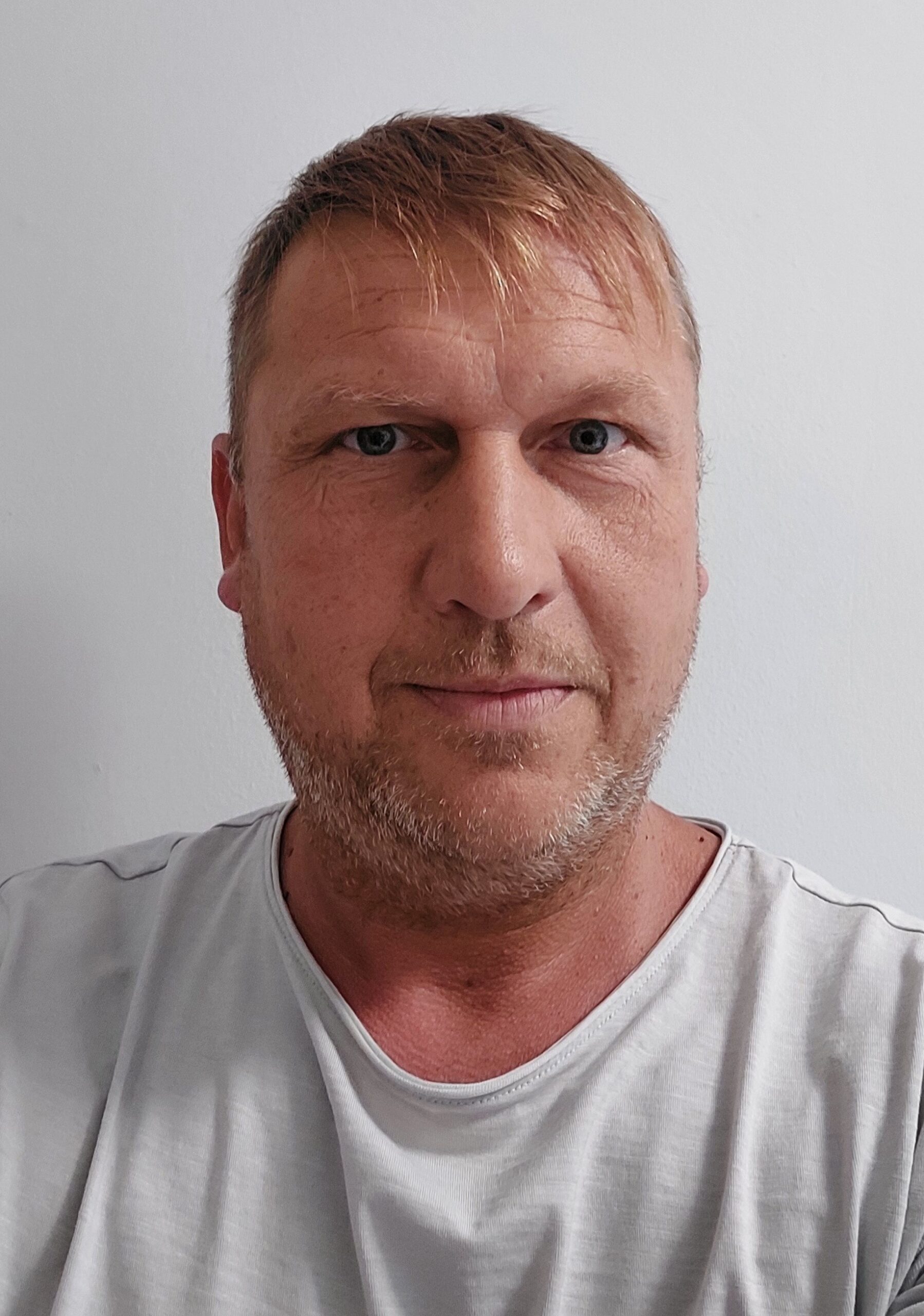 Ein Mann mittleren Alters mit kurzen hellbraunen Haaren und blauen Augen, der ein hellgraues T-Shirt trägt, steht vor einer schlichten weißen Wand und blickt mit neutralem Blick direkt in die Kamera, als würde er auf die Öffnungszeiten von Annahmestellen warten.