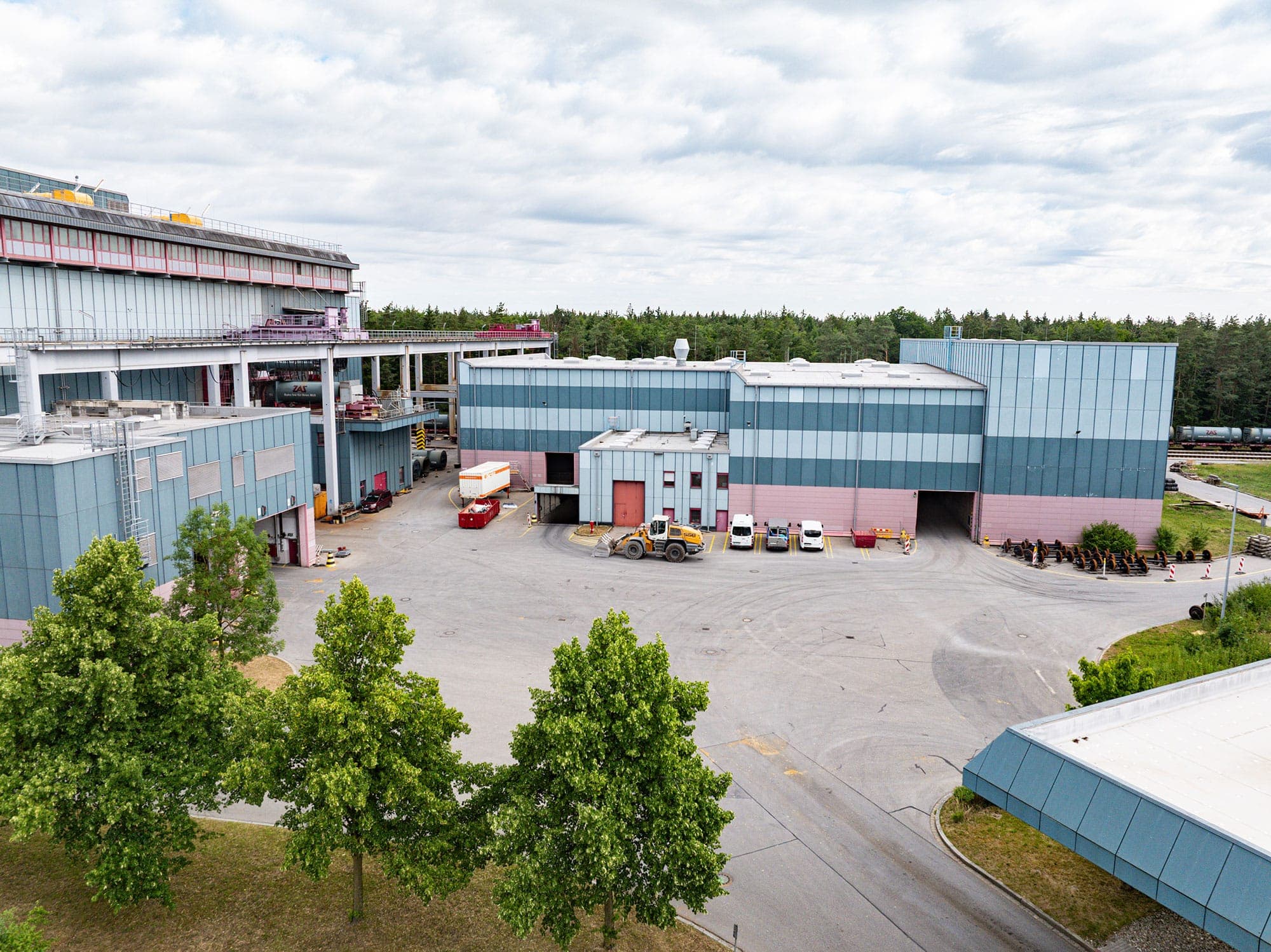 Eine Industrieanlage mit großen blauen und rosafarbenen Gebäuden, mehreren Fahrzeugen und Geräten auf dem Hof, umgeben von Bäumen und Grünflächen unter einem bewölkten Himmel.