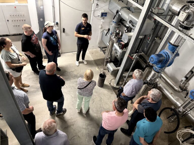 Eine Gruppe von Menschen steht im Kreis um Herrn Balz, der einen Vortrag in einer Industrieanlage hält, in der Rohre, Messgeräte und Maschinen an den Wänden zu sehen sind - zur Feier von 25 Jahren im Einsatz.