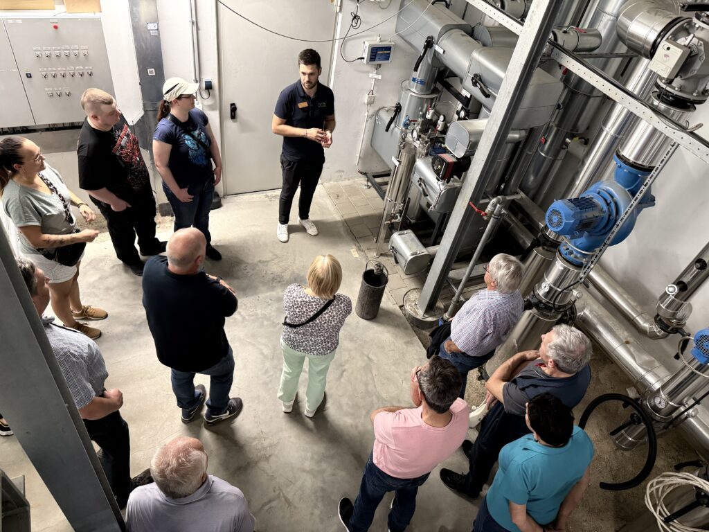 Eine Gruppe von Menschen steht im Kreis um Herrn Balz, der einen Vortrag in einer Industrieanlage hält, in der Rohre, Messgeräte und Maschinen an den Wänden zu sehen sind - zur Feier von 25 Jahren im Einsatz.