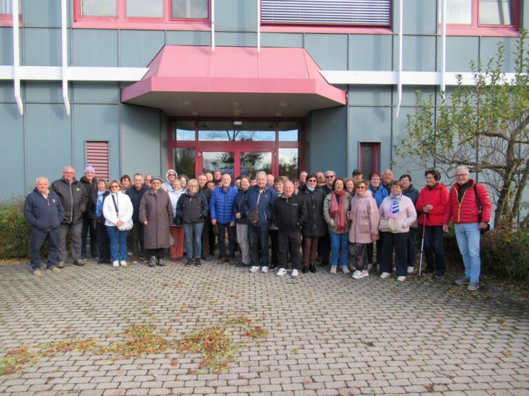 Eine große Gruppe von Menschen in Jacken und Mänteln steht zusammen und lächelt für ein Foto vor einem modernen Gebäude mit einem roten Eingangsvordach und großen Fenstern. Im Vordergrund sind Sträucher und gepflasterte Steine zu sehen.