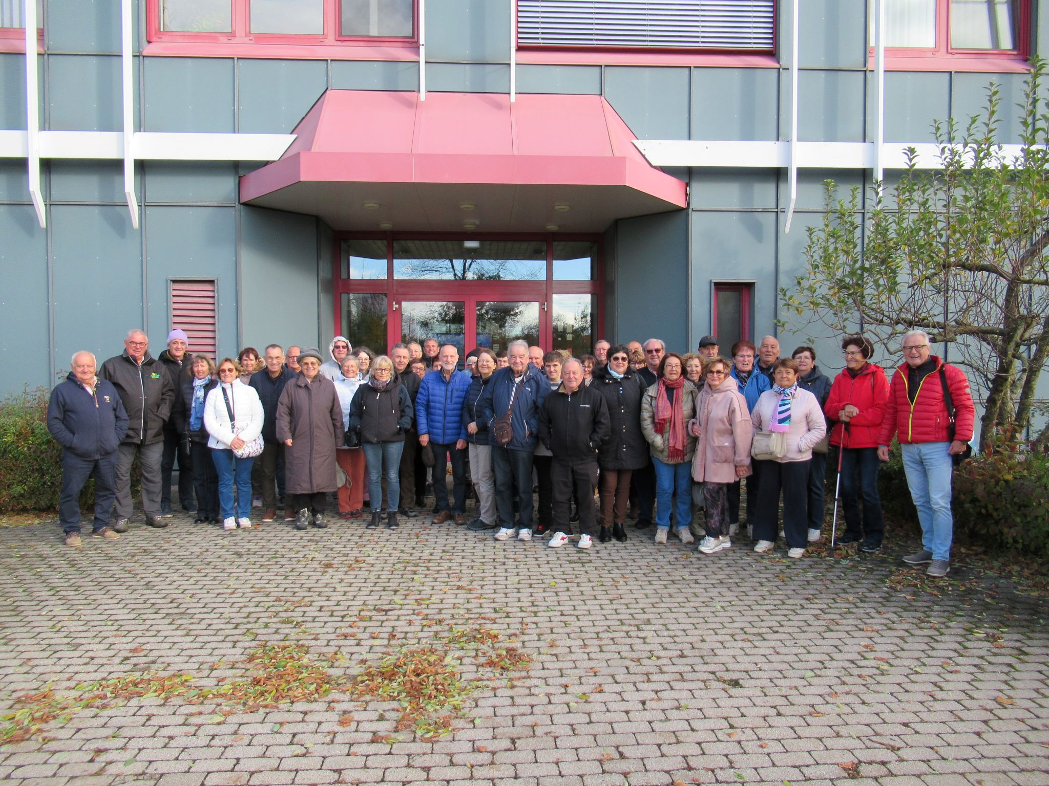 Eine große Gruppe von Menschen in Jacken und Mänteln steht zusammen und lächelt für ein Foto vor einem modernen Gebäude mit einem roten Eingangsvordach und großen Fenstern. Im Vordergrund sind Sträucher und gepflasterte Steine zu sehen.