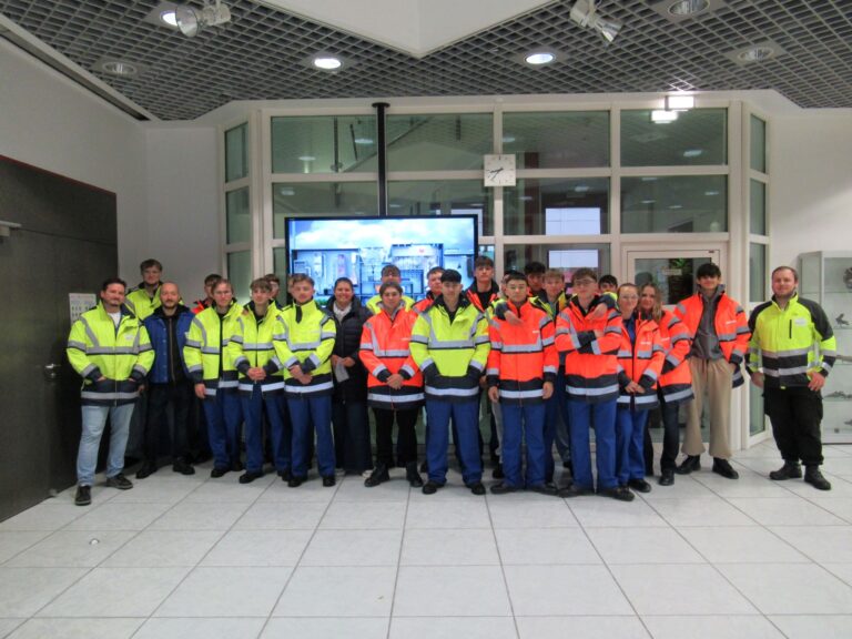 In einem hell erleuchteten Innenraum mit gefliesten Böden und Glaswänden steht eine Gruppe von Menschen, die meist Warnwesten in Gelb und Orange tragen, zusammen und posiert für ein Gruppenfoto.