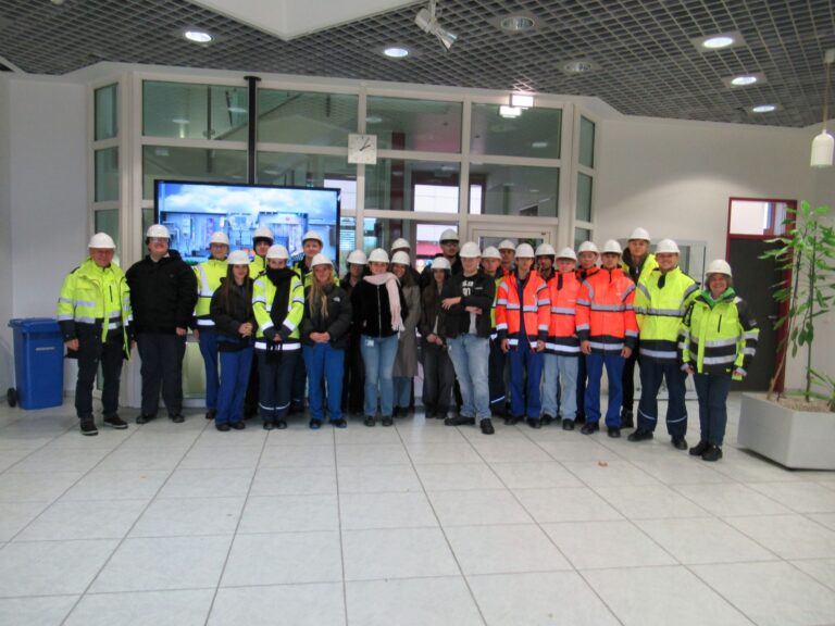 Eine Gruppe von Personen, die Schutzhelme und Warnwesten tragen, steht in einer modernen Büro- oder Industriehalle zusammen und posiert für ein Gruppenfoto.