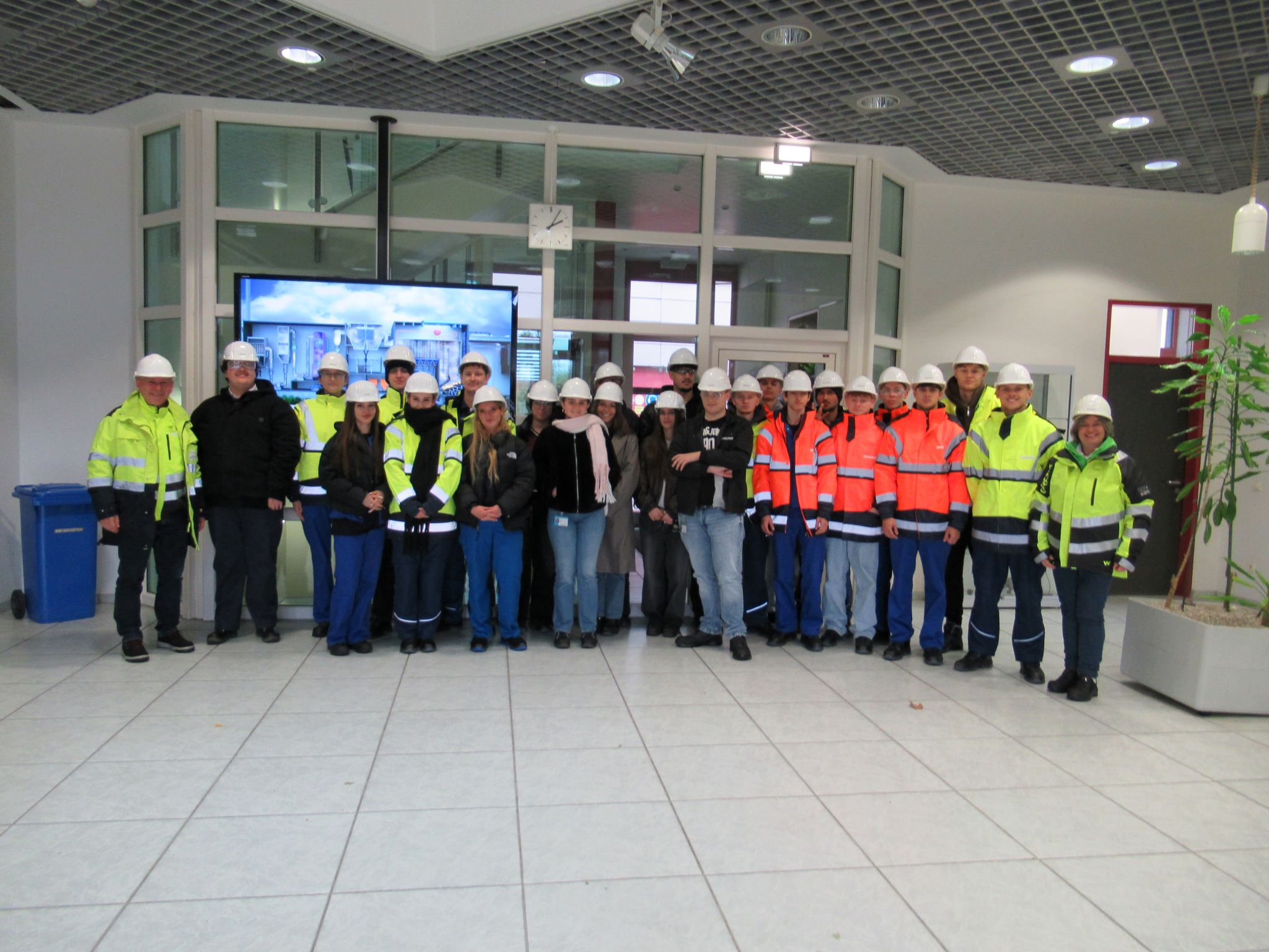 Eine Gruppe von Personen, die Schutzhelme und Warnwesten tragen, steht in einer modernen Büro- oder Industriehalle zusammen und posiert für ein Gruppenfoto.