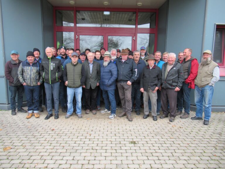 Eine Gruppe von etwa 30 Männern unterschiedlichen Alters steht dicht beieinander auf einem gepflasterten Platz vor einem modernen Gebäude mit großen Glastüren und Fenstern und posiert für ein Gruppenfoto.