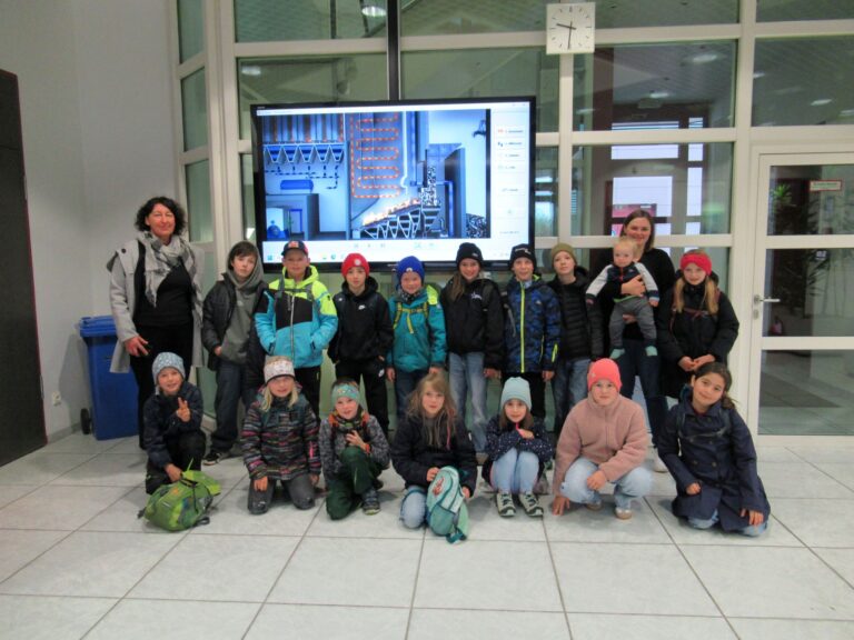 Eine Gruppe von Kindern und zwei Erwachsene posieren in einem Raum vor einem großen Bildschirm, auf dem Industriemaschinen zu sehen sind. Die meisten Kinder tragen Jacken und Hüte, und einige sitzen auf dem Boden, während andere hinter ihnen stehen.