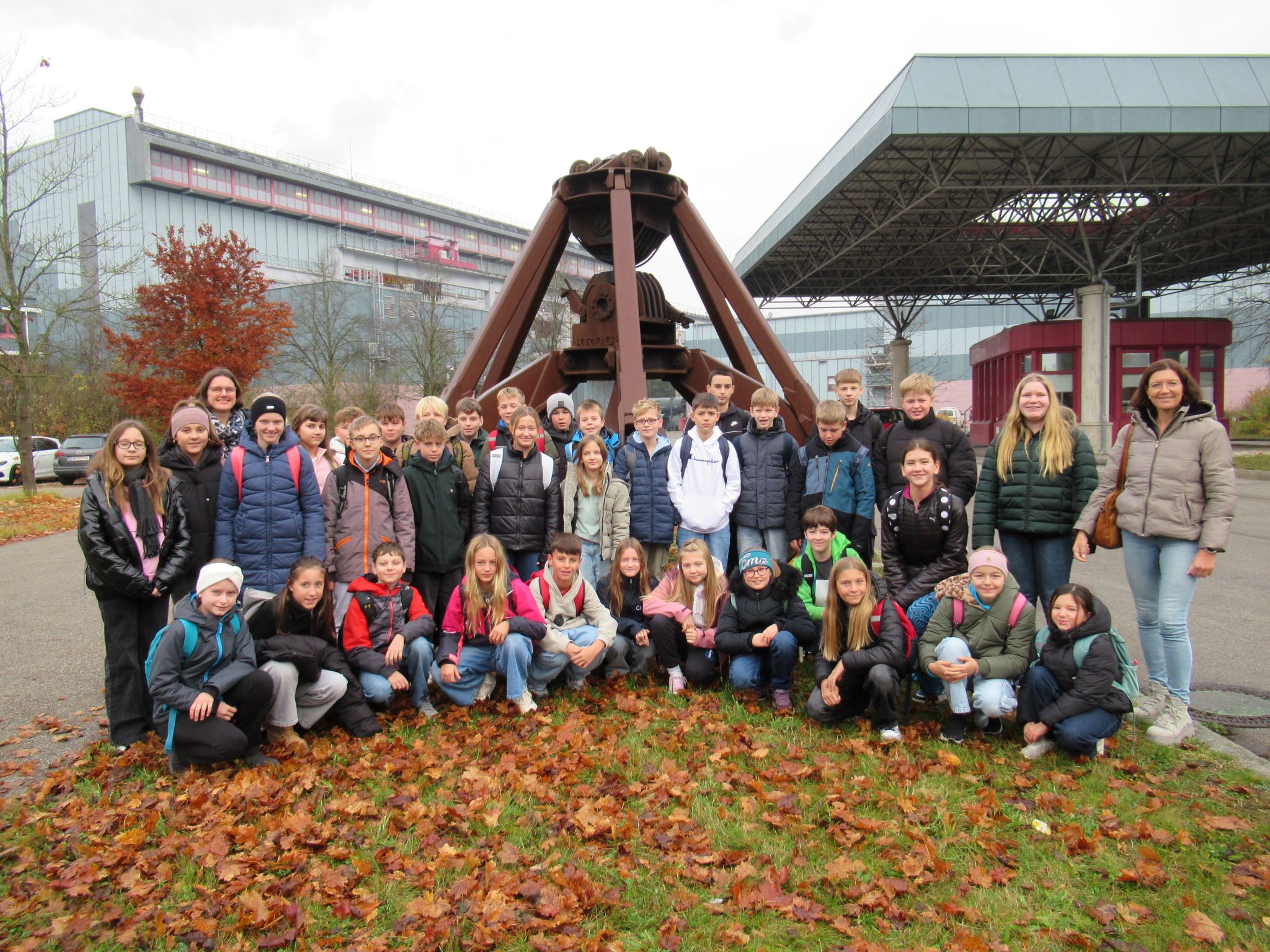 Eine Gruppe von Kindern und ein paar Erwachsene posieren gemeinsam im Freien vor einer großen Holzkonstruktion, deren Gras mit Herbstblättern bedeckt ist. Im Hintergrund sind moderne Gebäude unter einem bewölkten Himmel zu sehen.