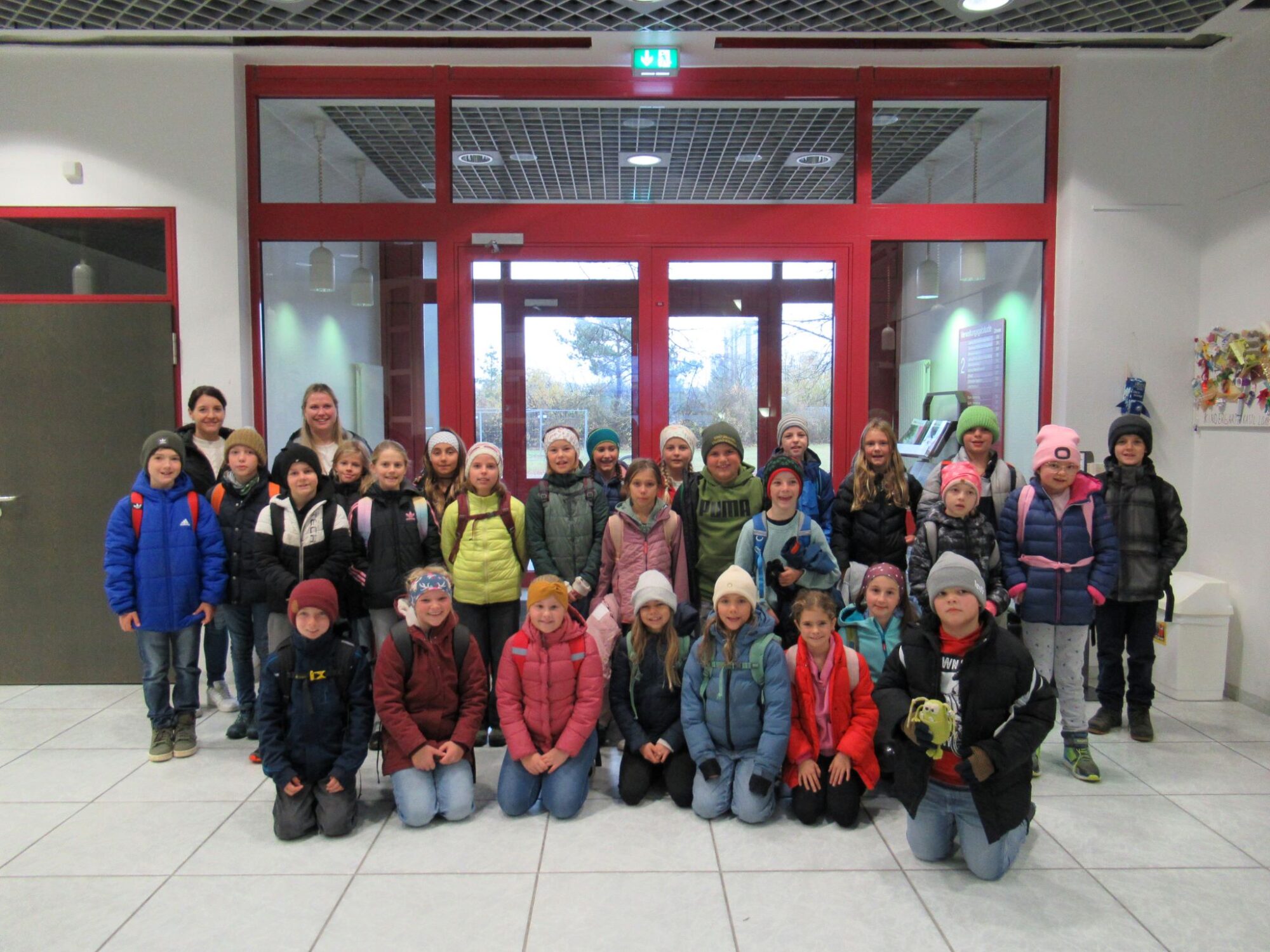 Eine Gruppe von Kindern in Winterjacken und M&uuml;tzen posiert f&uuml;r ein Foto in einem Innenraum vor einer Glast&uuml;r, in der hinteren Reihe stehen zwei Erwachsene. Die Kinder l&auml;cheln und scheinen bereit f&uuml;r einen Ausflug zu sein.