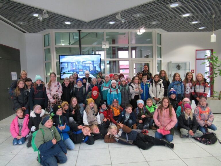 Eine große Gruppe von Kindern und einige Erwachsene posieren für ein Foto in einem hellen, modernen Eingangsbereich, mit Rucksäcken und Winterkleidung. Im Hintergrund sind ein großer Bildschirm und Glastüren zu sehen.