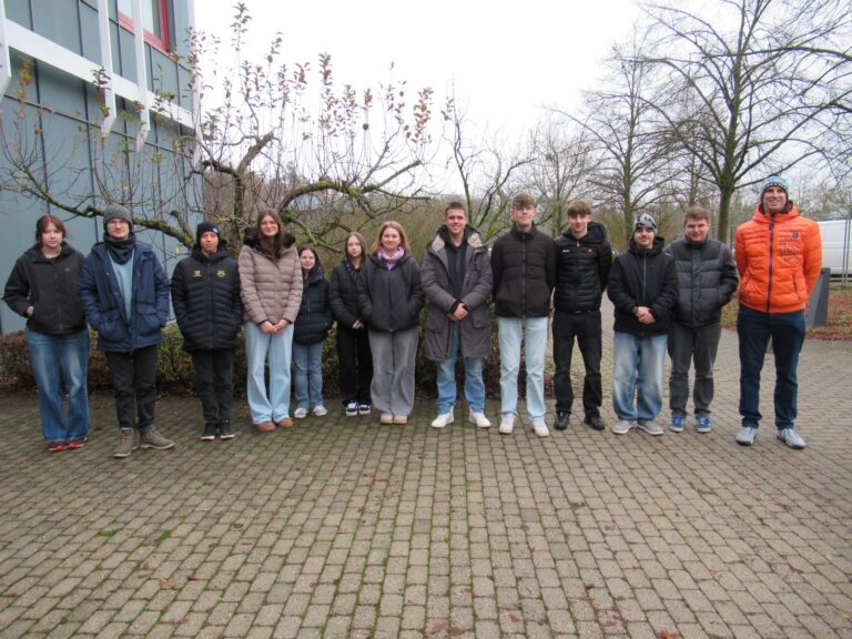 Eine Gruppe von zw&ouml;lf Personen in Winterjacken und Jeans steht in einer Reihe nebeneinander auf einem gepflasterten Platz mit kahlen B&auml;umen und einem Geb&auml;ude im Hintergrund.