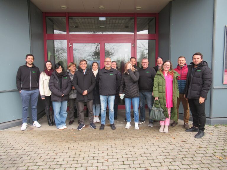 Eine Gruppe von fünfzehn Mitarbeitern der egis Verwaltungs GmbH steht lächelnd vor einem Gebäude mit großen rot gerahmten Glastüren. Die meisten sind in dunkle Jacken gekleidet, einige wenige tragen hellere Farben und stehen gemeinsam auf einem gepflasterten Außengelände.