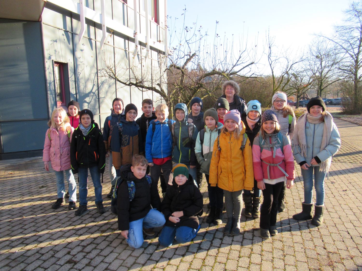 Eine Gruppe von Kindern und zwei Erwachsenen steht drau&szlig;en auf einem gepflasterten Platz in der N&auml;he eines Geb&auml;udes, warm gekleidet in Jacken, M&uuml;tzen und Schals, und l&auml;chelt an einem sonnigen Tag in die Kamera.