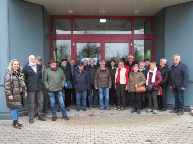 Eine Gruppe von etwa 20 Erwachsenen in Winterkleidung steht l&auml;chelnd vor einem Geb&auml;ude mit gro&szlig;en Glast&uuml;ren und roten Rahmen und posiert f&uuml;r ein Gruppenfoto auf einer gepflasterten Fl&auml;che.
