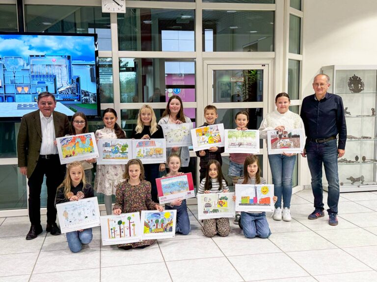 Eine Gruppe von Kindern mit bunten Zeichnungen posiert mit zwei Erwachsenen in einem hellen Raum mit gro&szlig;en Fenstern und einer Glasvitrine w&auml;hrend einer ZAS-Verbandsversammlung. Im Hintergrund sind ein digitaler Bildschirm und modernes Dekor zu sehen.