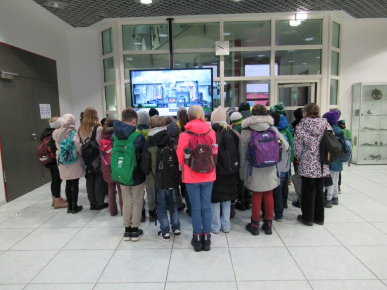 Eine Gruppe von Kindern mit M&auml;nteln und Rucks&auml;cken steht dicht beieinander und blickt auf einen gro&szlig;en Bildschirm in einem modernen Innenraum mit wei&szlig;en W&auml;nden, gefliestem Boden und Glasfenstern. Ihre R&uuml;cken sind der Kamera zugewandt.