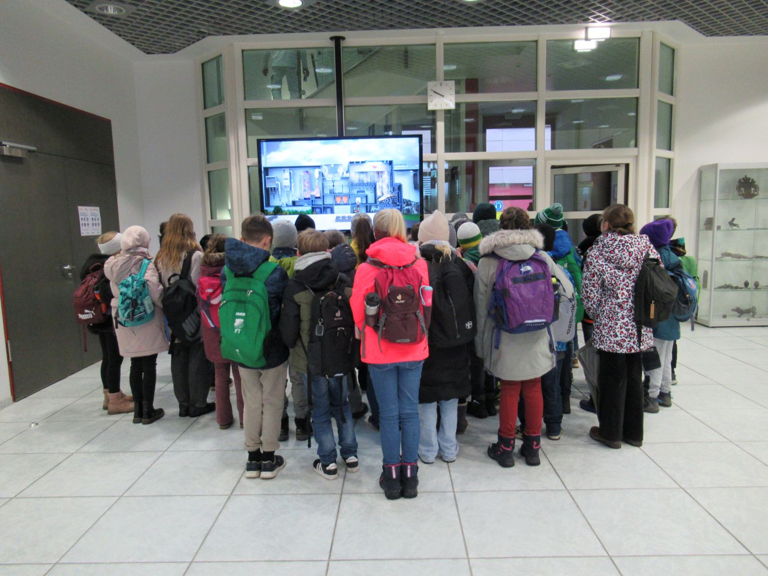 Eine Gruppe von Kindern mit M&auml;nteln und Rucks&auml;cken steht dicht beieinander und blickt auf einen gro&szlig;en Bildschirm in einem modernen Innenraum mit wei&szlig;en W&auml;nden, gefliestem Boden und Glasfenstern. Ihre R&uuml;cken sind der Kamera zugewandt.