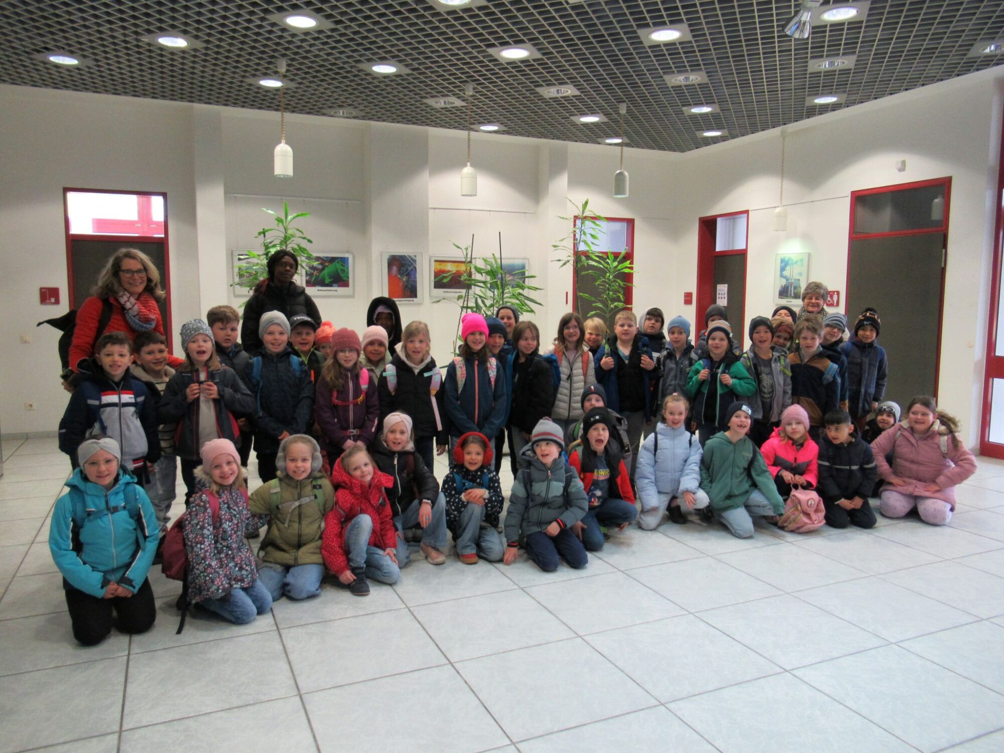 Eine gro&szlig;e Gruppe von Kindern aus der Grundschule Triftern und ein paar Erwachsene stellen sich f&uuml;r ein Gruppenfoto in einer Halle auf. Bunte Jacken und H&uuml;te erhellen die Szene vor wei&szlig;en W&auml;nden mit Kunstwerken, Pflanzen, einem gefliesten Boden und einer Decke mit Gittermuster im Jahr 2026.