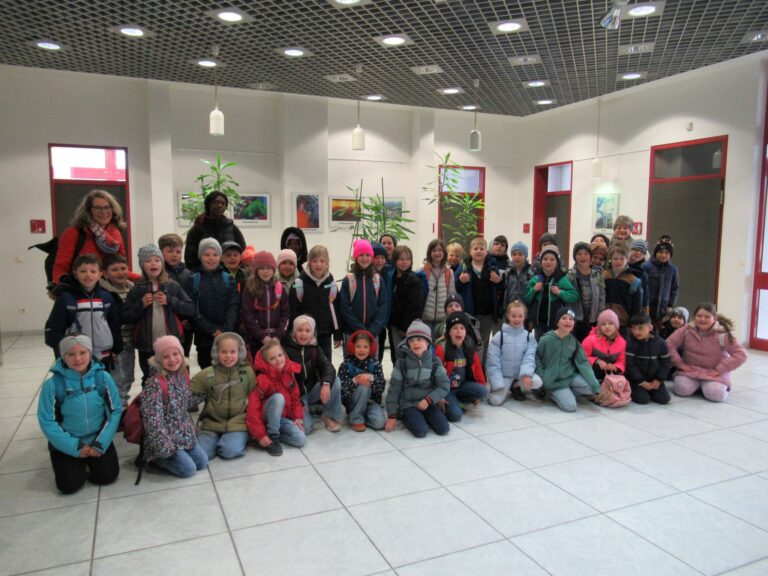 Eine gro&szlig;e Gruppe von Kindern aus der Grundschule Triftern und ein paar Erwachsene stellen sich f&uuml;r ein Gruppenfoto in einer Halle auf. Bunte Jacken und H&uuml;te erhellen die Szene vor wei&szlig;en W&auml;nden mit Kunstwerken, Pflanzen, einem gefliesten Boden und einer Decke mit Gittermuster im Jahr 2026.