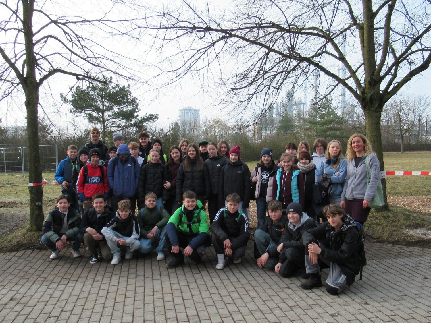 Eine Gruppe von Sch&uuml;lern und ein Erwachsener posieren im Freien auf einer gepflasterten Fl&auml;che, mit B&auml;umen und Gras im Hintergrund. Die meisten tragen Winterjacken und Rucks&auml;cke. Der Himmel ist bew&ouml;lkt und hinter ihnen befindet sich eine rot-wei&szlig;e Barriere.