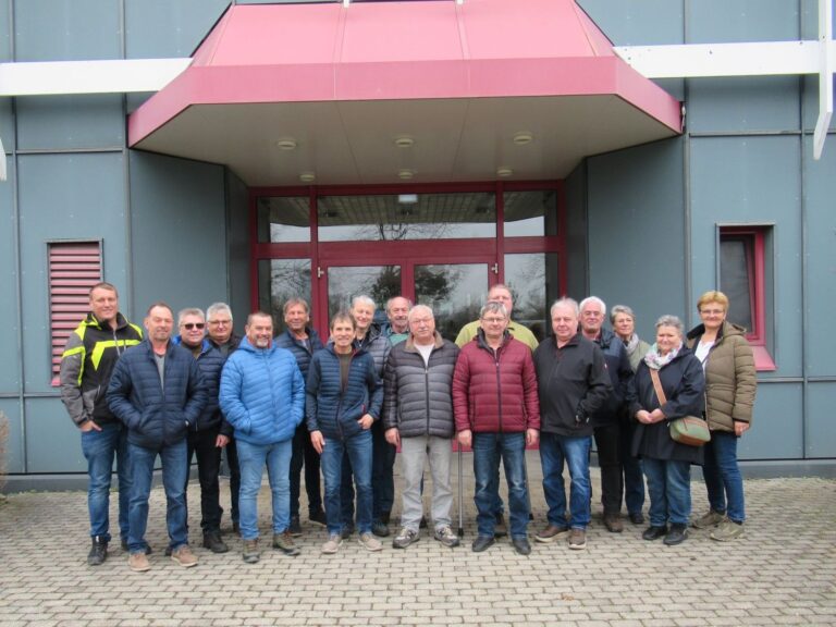 Eine Gruppe von sechzehn Erwachsenen steht in zwei Reihen vor dem Eingang eines modernen Geb&auml;udes, die meisten tragen Jacken und Jeans. Im Hintergrund sind eine Glast&uuml;r und graue W&auml;nde mit roten Akzenten zu sehen.
