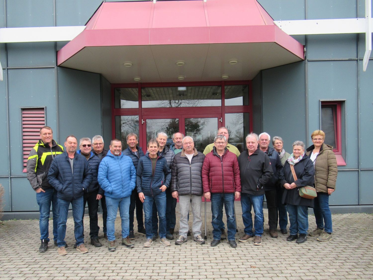Eine Gruppe von sechzehn Erwachsenen steht in zwei Reihen vor dem Eingang eines modernen Geb&auml;udes, die meisten tragen Jacken und Jeans. Im Hintergrund sind eine Glast&uuml;r und graue W&auml;nde mit roten Akzenten zu sehen.