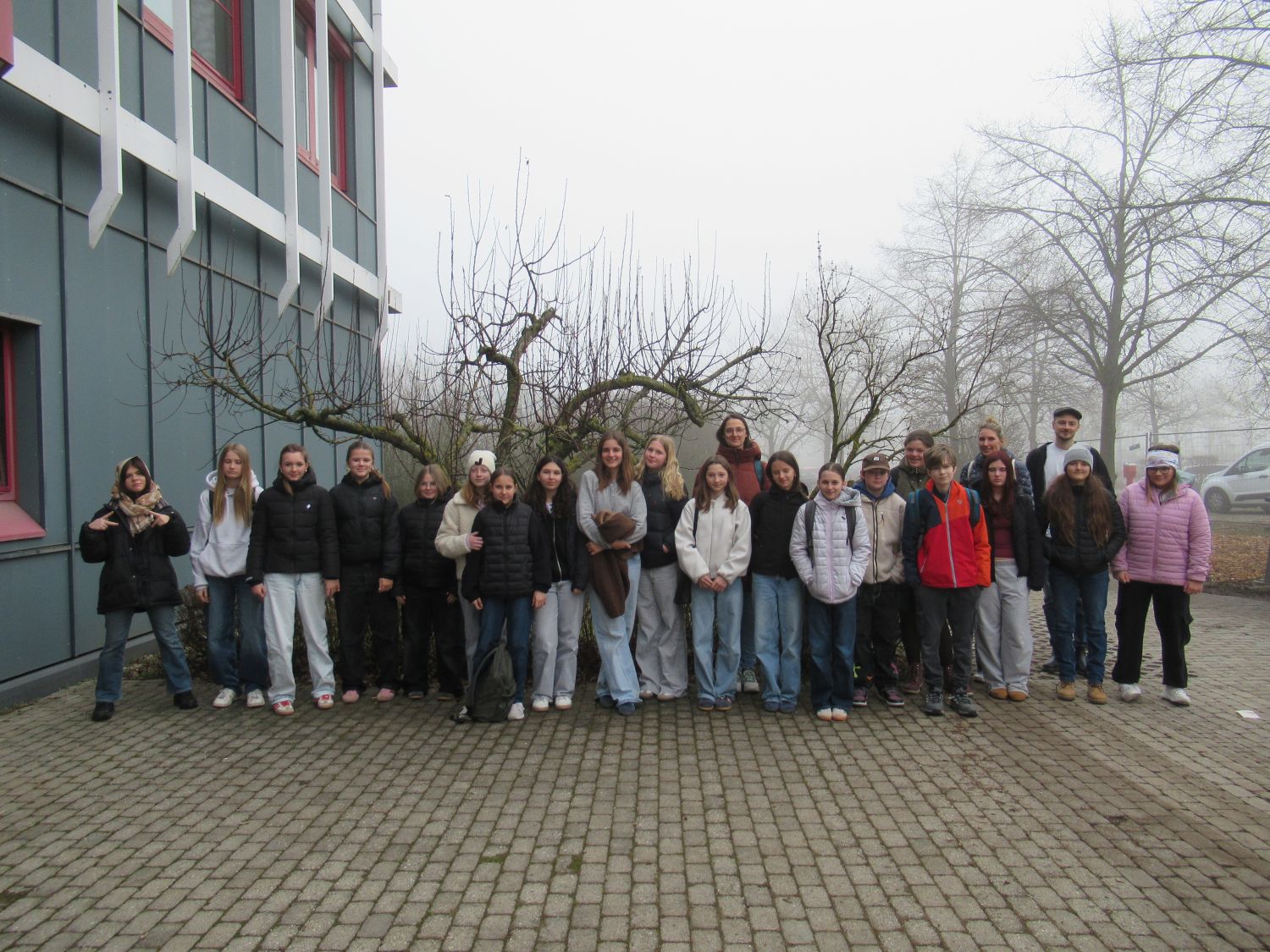 Eine Gruppe von etwa 24 Personen, meist Jugendliche und einige Erwachsene, steht an einem nebligen Tag auf einem gepflasterten Platz vor einem grauen Geb&auml;ude und blattlosen B&auml;umen und stellt sich f&uuml;r ein Gruppenfoto auf.