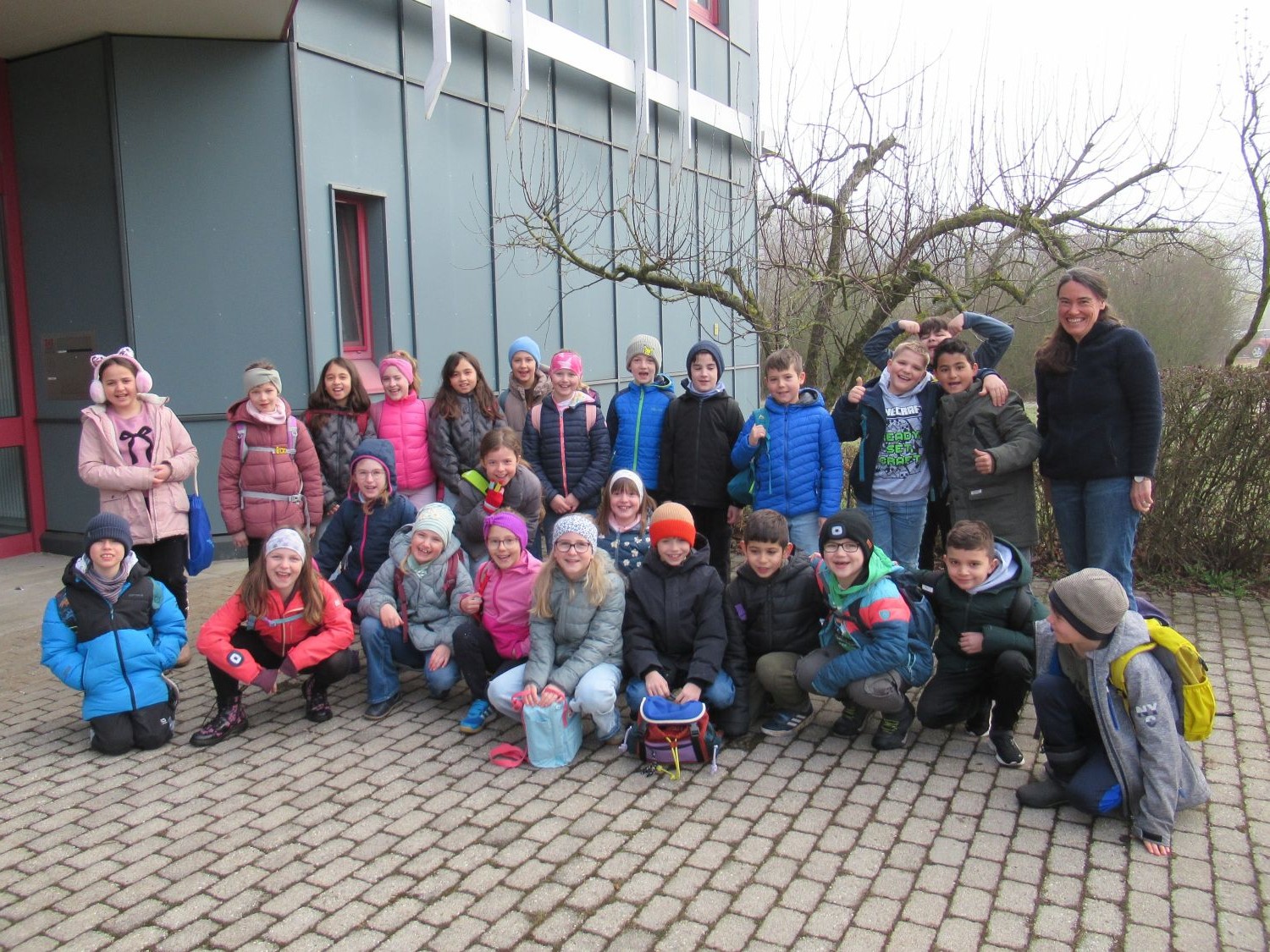 Eine Gruppe von Schulkindern und eine Frau stehen und knien gemeinsam vor einem modernen Geb&auml;ude auf einer gepflasterten Fl&auml;che. Die Kinder tragen bunte Winterkleidung, M&uuml;tzen und M&auml;ntel, und viele l&auml;cheln in die Kamera.