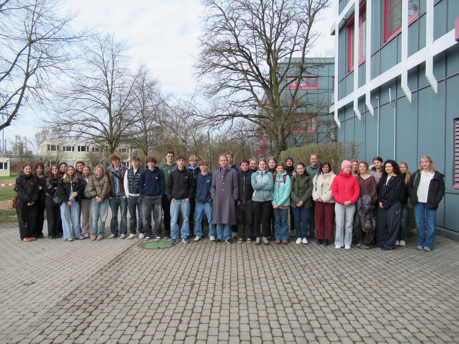 Eine gro&szlig;e Gruppe von Menschen steht in einer Reihe im Freien auf einer gepflasterten Fl&auml;che neben einem modernen Geb&auml;ude, wobei im Hintergrund blattlose B&auml;ume und weitere Geb&auml;ude zu sehen sind. Die Gruppe ist leger und f&uuml;r k&uuml;hles Wetter gekleidet.