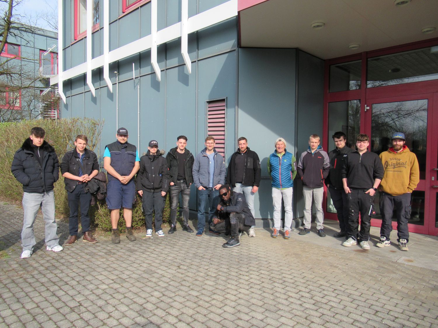 Dreizehn Personen stehen in einer Reihe vor einem modernen Geb&auml;ude mit grauen Paneelen und roten Fensterrahmen und posieren an einem sonnigen Tag f&uuml;r ein Gruppenfoto. Die meisten sind leger gekleidet, und eine Person hockt davor.