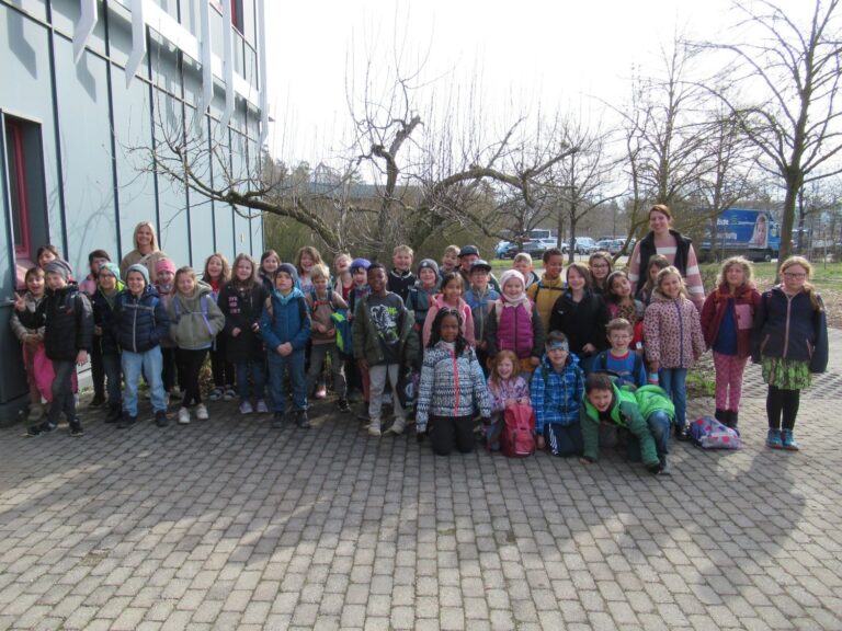 Eine gro&szlig;e Gruppe von Kindern und zwei Erwachsene posieren f&uuml;r ein Foto auf einer gepflasterten Fl&auml;che in der N&auml;he eines Geb&auml;udes, mit kahlen B&auml;umen und einem Parkplatz im Hintergrund.