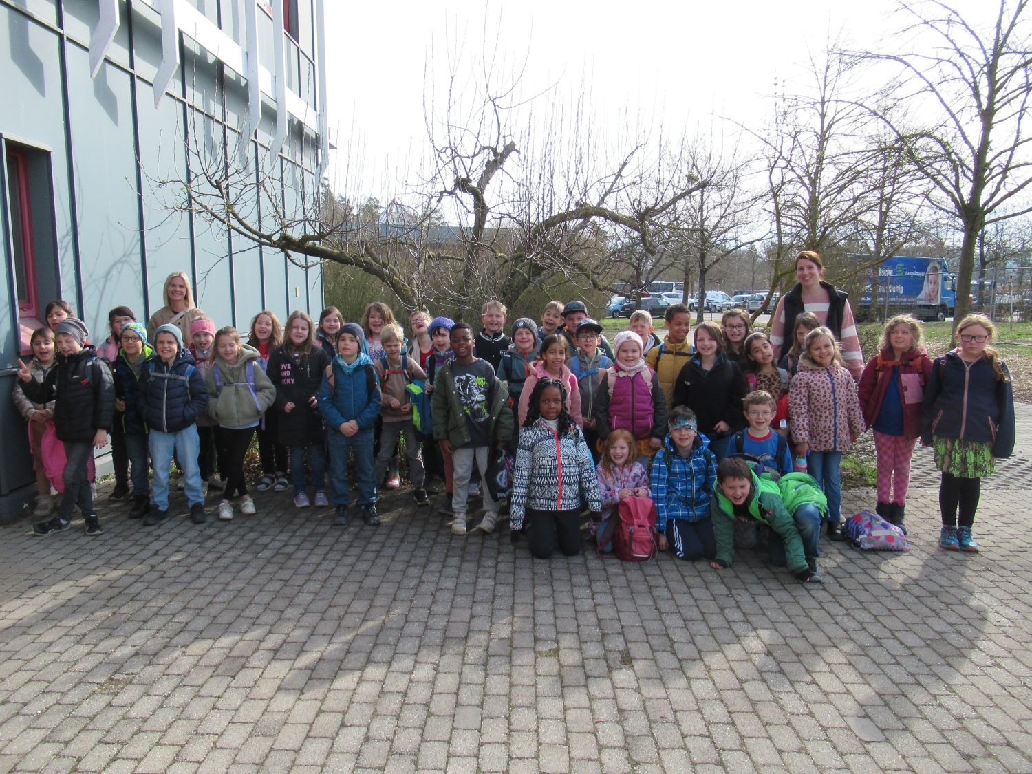 Eine gro&szlig;e Gruppe von Kindern und zwei Erwachsene posieren f&uuml;r ein Foto auf einer gepflasterten Fl&auml;che in der N&auml;he eines Geb&auml;udes, mit kahlen B&auml;umen und einem Parkplatz im Hintergrund.