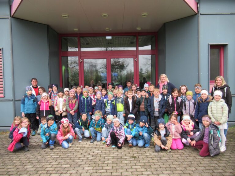 Eine gro&szlig;e Gruppe von Kindern und mehrere Erwachsene posieren und l&auml;cheln f&uuml;r ein Foto vor einem Geb&auml;ude mit Glast&uuml;ren. Die Kinder tragen bunte Jacken und H&uuml;te, und die Erwachsenen stehen an den Seiten und im Hintergrund.