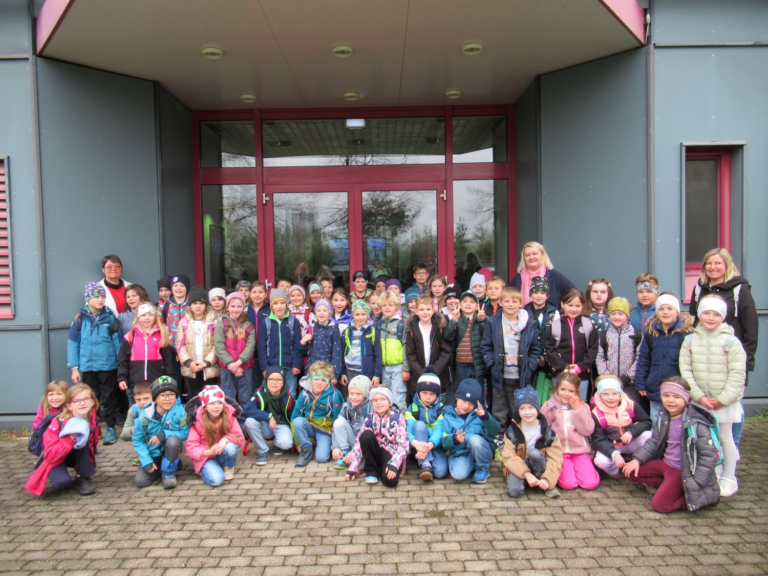 Eine gro&szlig;e Gruppe von Kindern und mehrere Erwachsene posieren und l&auml;cheln f&uuml;r ein Foto vor einem Geb&auml;ude mit Glast&uuml;ren. Die Kinder tragen bunte Jacken und H&uuml;te, und die Erwachsenen stehen an den Seiten und im Hintergrund.