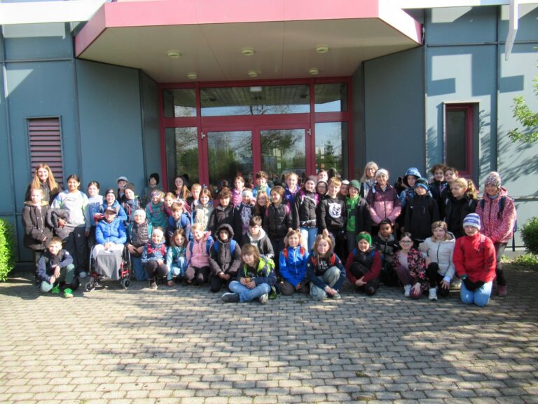 Eine gro&szlig;e Gruppe von Kindern und einige Erwachsene posieren f&uuml;r ein Foto vor einem Geb&auml;ude mit Glast&uuml;ren, stehend und sitzend auf einer gepflasterten Fl&auml;che, bekleidet mit Jacken und Freizeitkleidung.