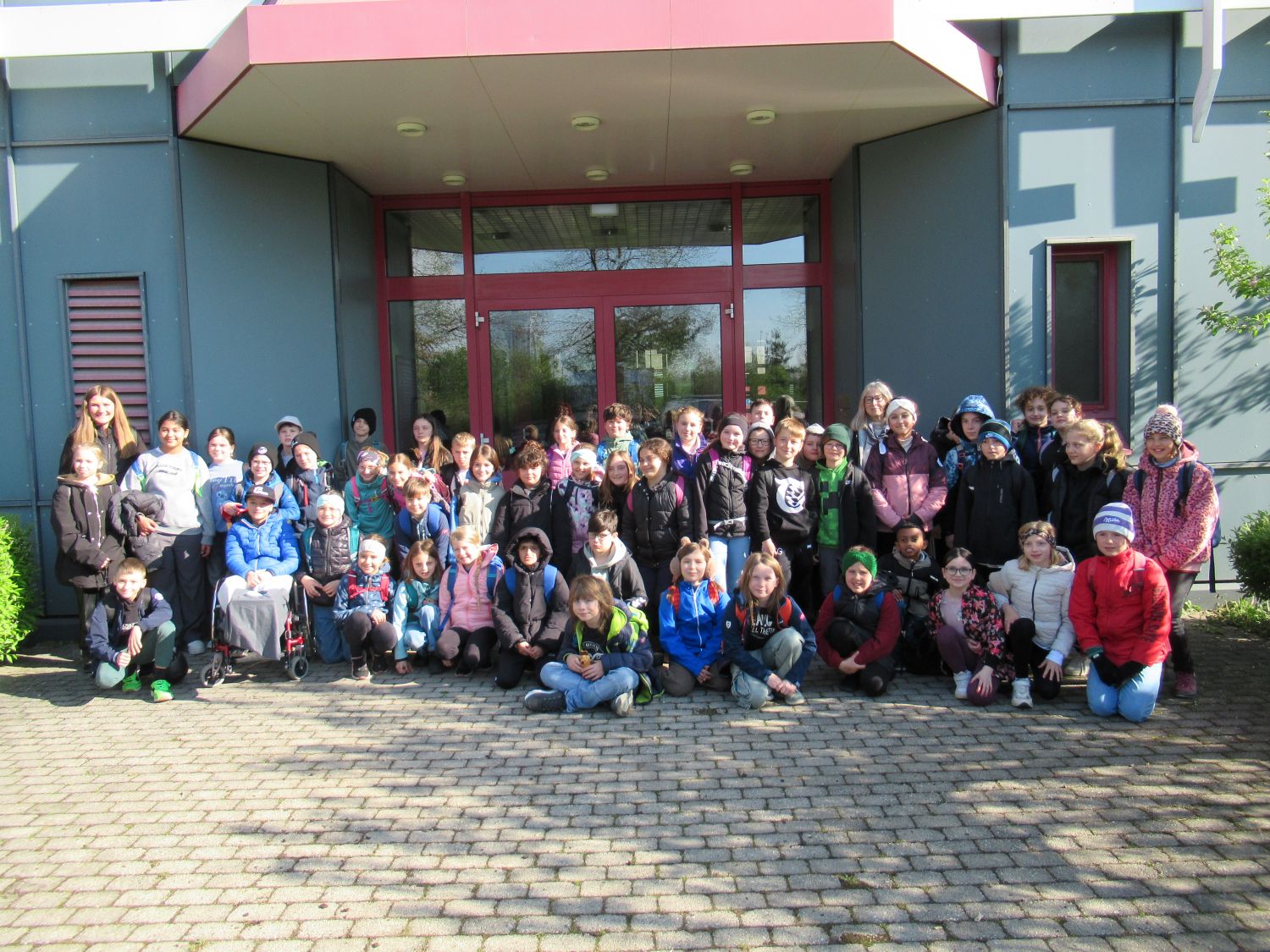 Eine gro&szlig;e Gruppe von Kindern und einige Erwachsene posieren f&uuml;r ein Foto vor einem Geb&auml;ude mit Glast&uuml;ren, stehend und sitzend auf einer gepflasterten Fl&auml;che, bekleidet mit Jacken und Freizeitkleidung.