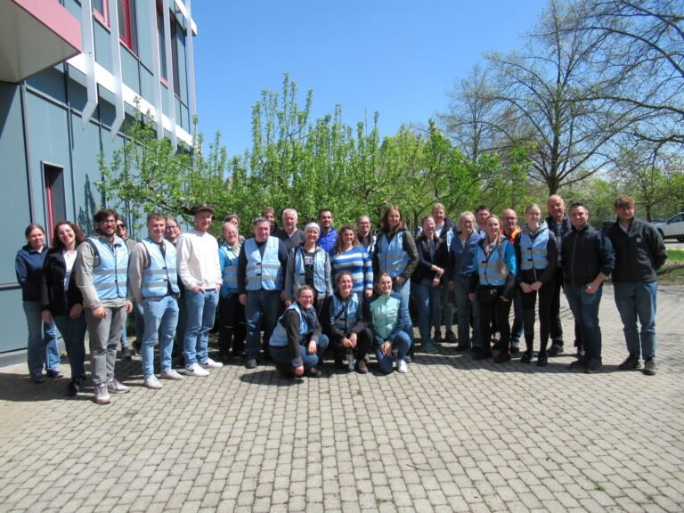 Eine Gruppe von etwa 30 Personen, von denen einige hellblaue Westen tragen, steht an einem sonnigen Tag vor einem Geb&auml;ude, mit gr&uuml;nen B&auml;umen und blauem Himmel im Hintergrund.