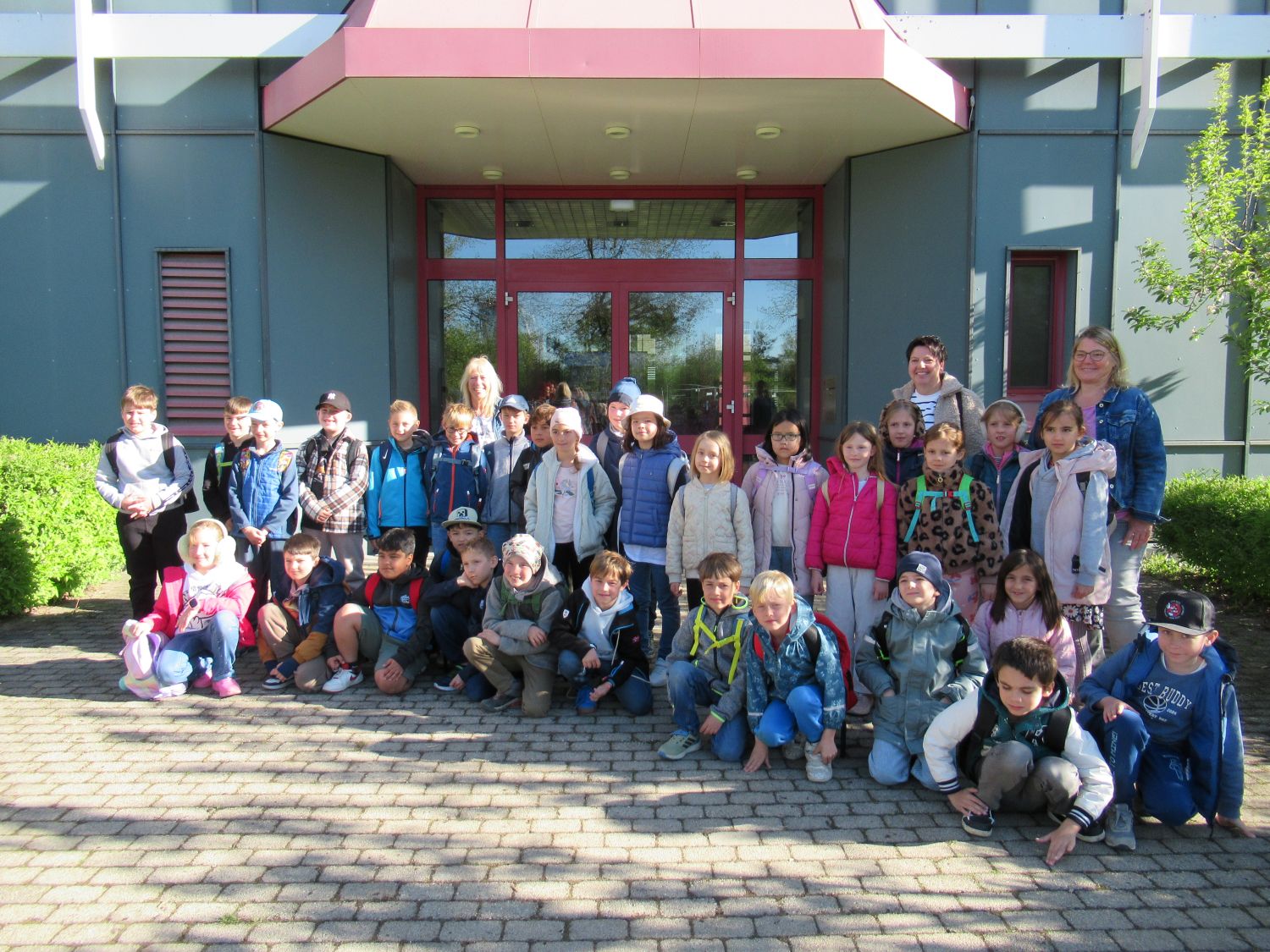 Eine Gruppe von Kindern und zwei Erwachsenen posieren gemeinsam vor einem Geb&auml;ude mit Glast&uuml;ren und roter Umrandung. Sie stehen und sitzen auf einer gepflasterten Fl&auml;che unter einem sonnigen Himmel.