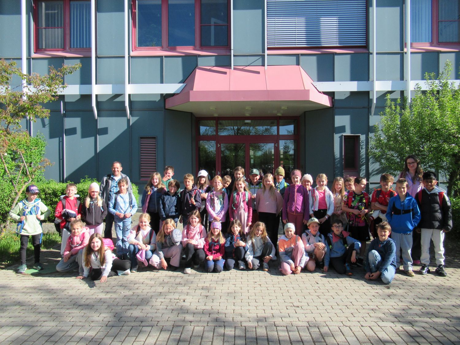 Eine gro&szlig;e Gruppe junger Kinder und ein Erwachsener stehen und sitzen an einem sonnigen Tag vor einem modernen Geb&auml;ude mit blauen W&auml;nden und roten Verzierungen und stellen sich f&uuml;r ein Gruppenfoto auf. Die meisten Kinder tragen Rucks&auml;cke und Freizeitkleidung.