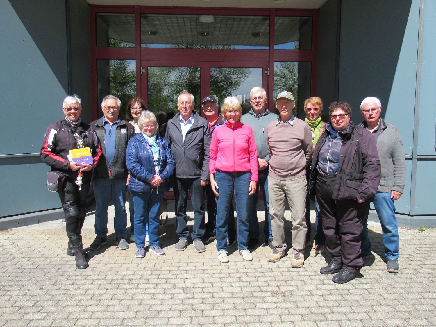 Eine Gruppe von dreizehn &auml;lteren Erwachsenen steht an einem sonnigen Tag auf einem gepflasterten Platz vor einem Geb&auml;ude mit gro&szlig;en Glast&uuml;ren zusammen und l&auml;chelt in die Kamera.