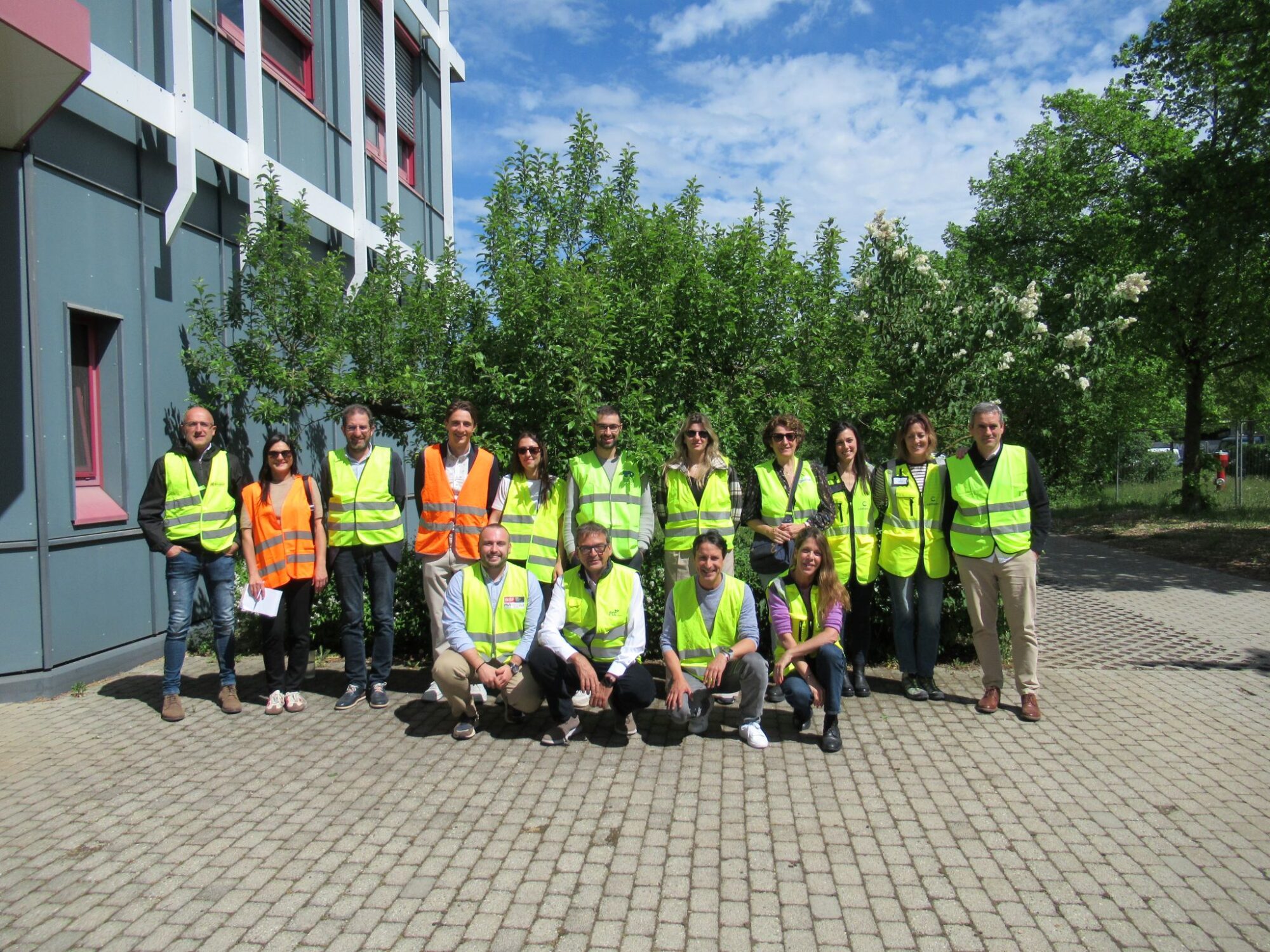 Eine Gruppe von Personen in gelben und orangefarbenen Sicherheitswesten, die ACLIMA und den Baskischen Umweltcluster repr&auml;sentieren, posiert im Freien auf einer gepflasterten Fl&auml;che vor einem Geb&auml;ude und gr&uuml;nen B&auml;umen unter einem teilweise bew&ouml;lkten Himmel, um ihre Zusammenarbeit im Jahr 2026 zu feiern.
