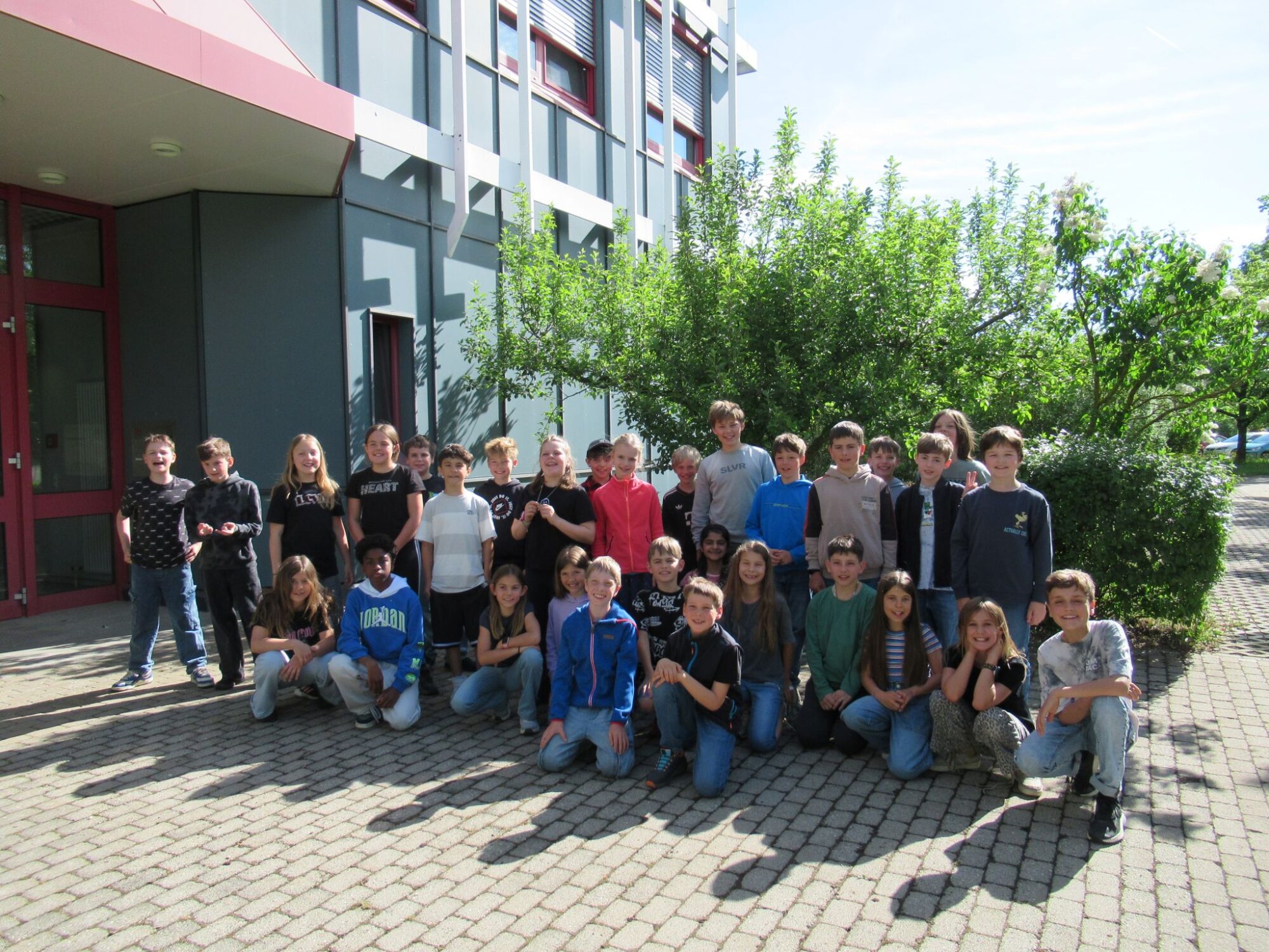 Eine Gruppe von Schulkindern der Grundschule Chieming posiert an einem sonnigen Tag f&uuml;r ein Foto vor einem Geb&auml;ude. Sie stehen und knien auf einem Steinweg mit Gr&uuml;n und B&auml;umen im Hintergrund.