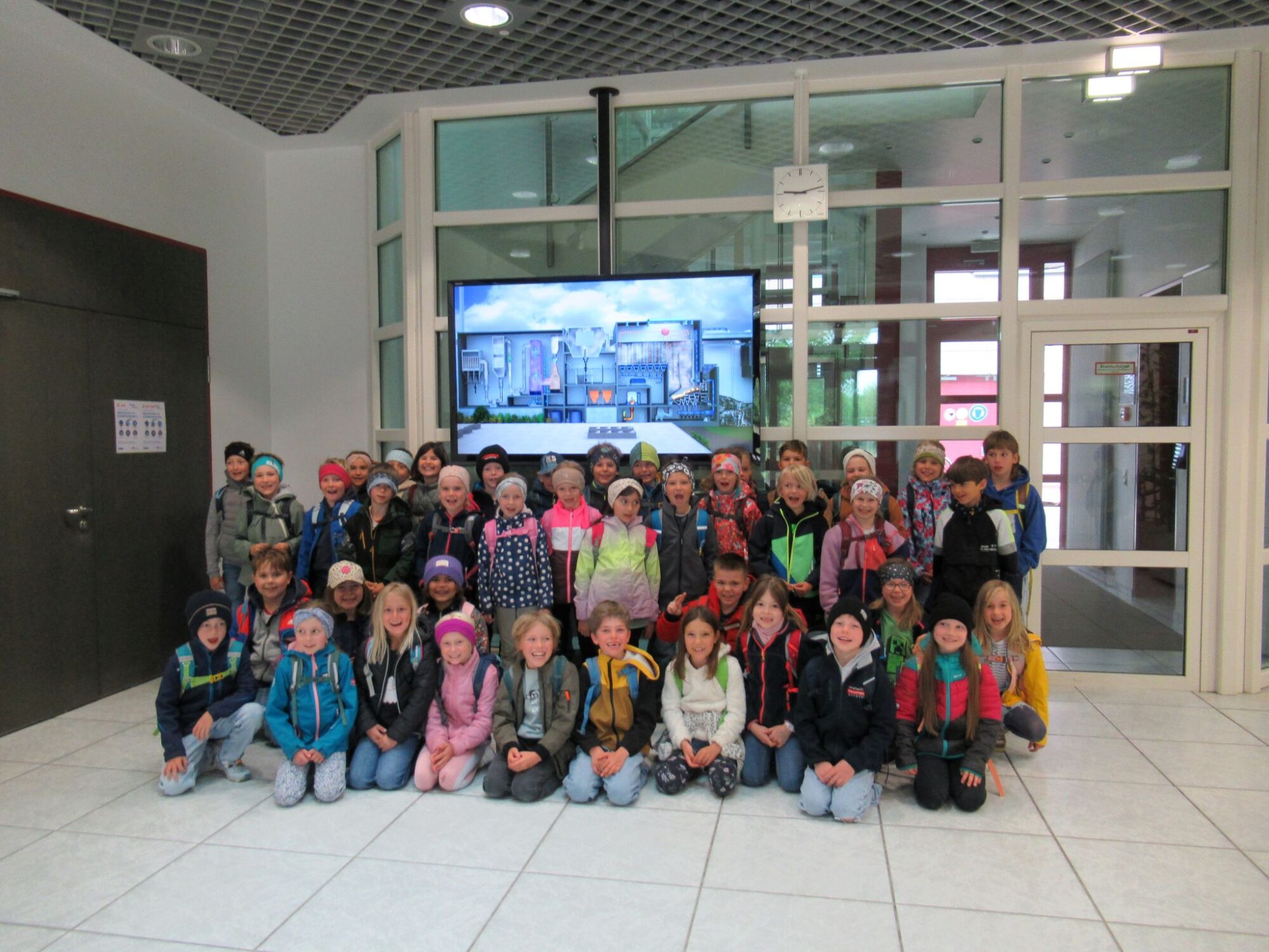 Eine gro&szlig;e Gruppe junger Kinder aus der Grundschule Grabenst&auml;tt, die Jacken und Rucks&auml;cke tragen, posieren f&uuml;r ein Gruppenfoto in einer Halle im Jahr 2026 vor einem an der Wand befestigten Bildschirm, auf dem ein Stadtbild zu sehen ist.